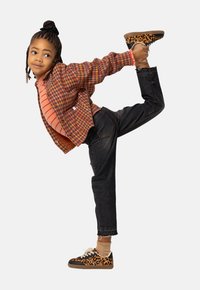 Child with braided hair stretching leg backward, wearing leopard-print shoes, dark jeans, striped shirt, and checkered jacket on white background.