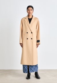 Cappotto doppiopetto in misto lana beige con grandi bottoni neri, rever a contrasto e polsino nero, indossato sopra una gonna di jeans.