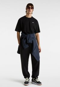 Schwarzes Baumwoll-T-Shirt mit Logo, dunkelblaues Hemd um die Taille gebunden, schwarze Jogginghosen und schwarze Sneakers mit weißen und braunen Akzenten.