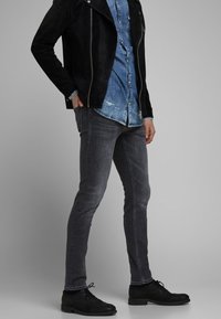 Schwarze Wildlederjacke, blaues Denimhemd und dunkelgraue Slim-Fit-Jeans mit schwarzen Schnürschuhen, die eine glatte Textur und minimale Details aufweisen.