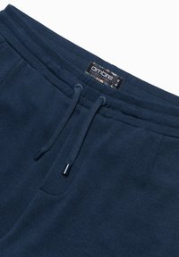 Tmavě modré tepláky vyrobené z texturované látky. Mají stažený pas se šňůrkou s plastovými konci a přišitým štítkem označeným "ombre."