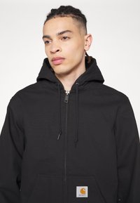 Black zip-up hoodie made of sturdy material, featuring a drawstring hood, front pocket, and a Carhartt logo tag.