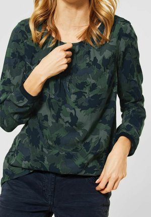 Femme portant un chemisier vert foncé à manches longues avec un motif de feuilles, tenant l'encolure d'une main et la hanche de l'autre.