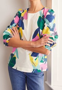 Person with arms crossed wearing a multicolored floral blouse with puffed sleeves and blue jeans, standing near a white wall.