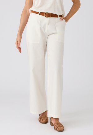 Mujer con pantalones blancos de talle alto y pierna ancha con bolsillos delanteros, cinturón marrón perforado y zapatos tejidos marrones claros con tacón bajo.