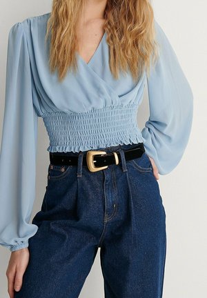 Woman wearing light blue smocked waist blouse with long puff sleeves, dark high-waisted jeans, and black belt with gold buckle.