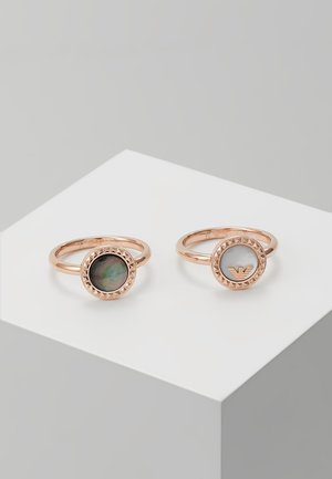 Twee roségouden ringen met ronde zettingen; links heeft een donkere iriserende steen, rechts heeft een witte steen met een gouden embleem erin.