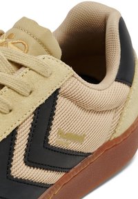 Scarpa sportiva beige con tomaia in tessuto texture, dettagli in pelle nera e suola in gomma marrone. Presenta un design cucito e dettagli con logo.