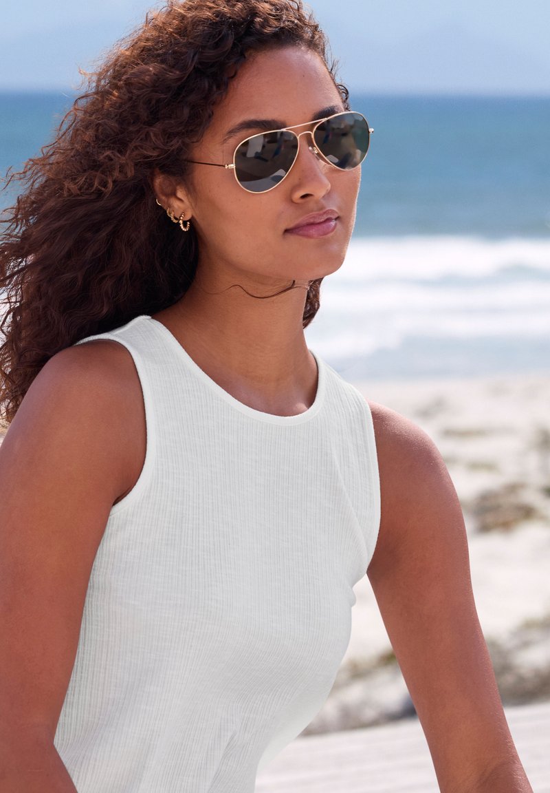 White sleeveless top with a textured fabric, paired with gold aviator sunglasses and hoop earrings, set against a beach background.