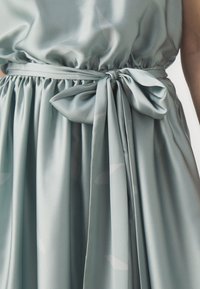 Λεπτομέρεια από γαλάζιο σατινένιο φόρεμα με μαζεμένη μέση και δεμένο φιόγκο. Ο ιστός είναι λείος με υπο subtle υφή και απαλή ρέουσα εφαρμογή.