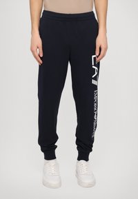 Marine joggebukser laget av mykt stoff, med elastisk midjebånd og mansjetter, og hvit logo trykt på venstre ben.