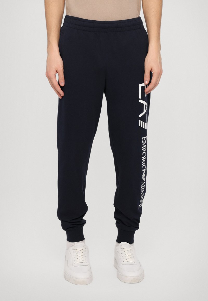 Marine joggebukser laget av mykt stoff, med elastisk midjebånd og mansjetter, og hvit logo trykt på venstre ben.