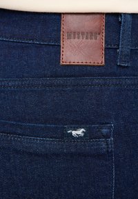 Ciemnoniebieskie dżinsy z denimu, wyposażone w brązową skórzaną naszywkę z napisem "MUSTANG" oraz małą niebieską metką z białym logo konia.