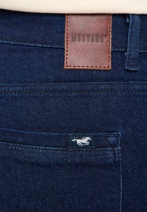 Ciemnoniebieskie dżinsy z denimu, wyposażone w brązową skórzaną naszywkę z napisem "MUSTANG" oraz małą niebieską metką z białym logo konia.