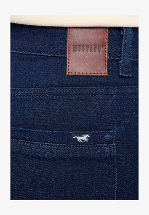 Ciemnoniebieskie dżinsy z denimu, wyposażone w brązową skórzaną naszywkę z napisem "MUSTANG" oraz małą niebieską metką z białym logo konia.