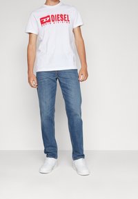 Camiseta blanca de algodón con estampado rojo "DIESEL", combinada con vaqueros de lavado medio y zapatillas blancas. Conjunto sencillo y casual en exhibición.