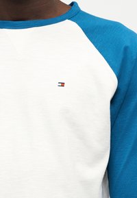 Κοντινό πλάνο ενός ατόμου που φοράει ένα λευκό πουκάμισο με μπλε raglan μανίκια και ένα μικρό κόκκινο, άσπρο και μπλε ορθογώνιο λογότυπο στο στήθος.