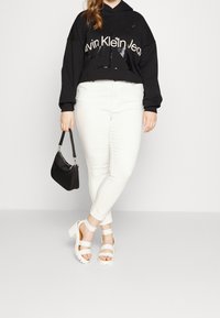 Czarna krótka bluza z kapturem z białym logo, białe jeansy slim-fit i białe sandały na obcasie. Czarna torba na krótkiem uchwycie. Prosty design, czyste kolory.