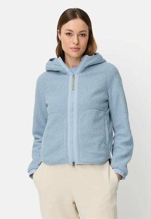 Kvinde iført lyseblå fleecejakke med hætte og lynlås, hænderne i lommerne, beige bukser, stående mod ensfarvet baggrund.