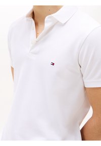 Bílé polo tričko z texturované látky, s tříknoflíkovou légou a malým vyšitým logem v červené, bílé a modré barvě.