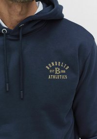 Mężczyzna w granatowej bluzie z kapturem z beżowym logo "Bondelid Athletics EST 1956" na klatce piersiowej i granatowymi sznurkami.