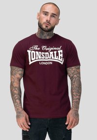 Tätoveeringutega mees kannab bordoopunast "Lonsdale London" t-särki ja musti rebenenud teksaseid halli ühtlase tausta ees.