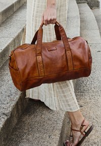 Borsa da viaggio in pelle marrone con doppi manici, chiusura con zip e tasca laterale, caratterizzata da una texture liscia e un sottile logo del marchio sul davanti.