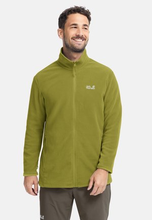 Lachende man die een olijfgroene fleecejas met ritssluiting en hoge kraag draagt, met het Jack Wolfskin-logo op de borst, staand tegen een effen achtergrond.