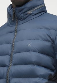 Vīrietis tērpies tumši zilā pūkainā jakā ar augstu apkakli, redzamu sudraba rāvējslēdzēju un CK logotipu uz krūtīm.