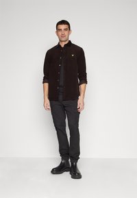 Camisa de pana negra con botones al frente, combinada con pantalones cargo negros. Botas negras hasta el tobillo con suela gruesa, de pie contra un fondo liso.