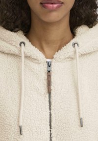 Kreemikas fliisist kapuutsiga sviiter, millel on tõmblukk, reguleeritav kapuuts, tekstureeritud kangas ja nahast aksessuaar tõmbluku küljes.