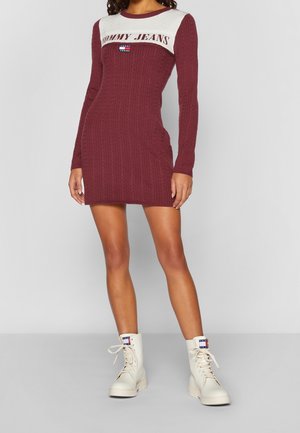Kobieta w bordowej dzianinowej sukience z napisem i logo "Tommy Jeans" na piersi, w połączeniu z białymi sznurowanymi botkami za kostkę z logo.