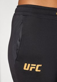 Svart trening korte med en gull "UFC" logo, med en jevn tekstur og et mesh-panel på siden for pusteevne.