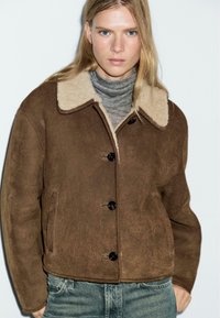 Bruine suède jas met een beige schapenvachtkraag, voorzien van zwarte knopen, zijzakken en een cropped ontwerp. Gedragen over een grijze coltrui.