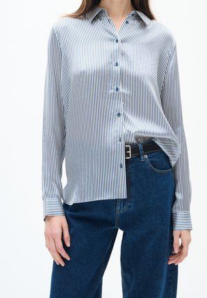 Woman wearing a blue and white vertical striped button-up shirt tucked into high-waisted dark blue jeans with a black belt.