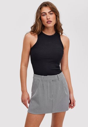 Jeune femme aux cheveux ondulés portant un haut noir sans manches et une jupe taille haute à carreaux noirs et blancs, debout devant un fond uni.