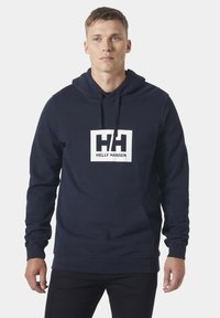 Sudadera de color azul marino hecha de algodón. Presenta un logo rectangular blanco con el texto "HH" y "HELLY HANSEN". Incluye un bolsillo tipo canguro y capucha con cordón.