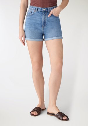 Donna con pantaloncini di jeans blu arrotolati, top marrone senza maniche e sandali scivolati marrone scuro, in piedi contro uno sfondo bianco semplice.