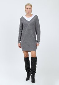 Grau gestricktes Kleid mit V-Ausschnitt, das diagonale Zopfmuster zeigt. Kombiniert mit hohen schwarzen Lederstiefeln, was einen mehrschichtigen Look schafft.