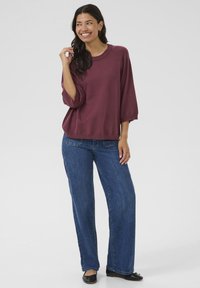 Maglietta morbida di colore bordeaux a maniche tre quarti, abbinata a jeans larghi blu. Il tessuto sembra soffice e ha una consistenza liscia. Ai piedi, ballerine nere.