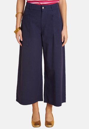 Woman wearing navy wide-leg cropped pants with front patch pockets, a striped pink top, gold bangles, and gold flat shoes.