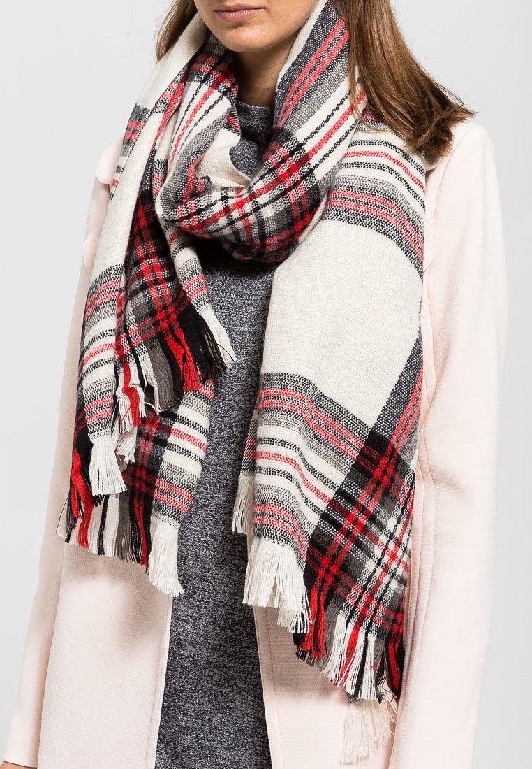 Femme portant une écharpe à carreaux rouge, noire et blanche avec des franges sur un haut gris et un manteau rose clair.