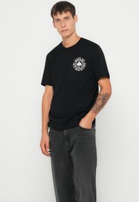Camiseta de algodón negra con cuello redondo, mangas cortas y un logo gráfico blanco en el pecho. Se lleva con jeans gris oscuro.