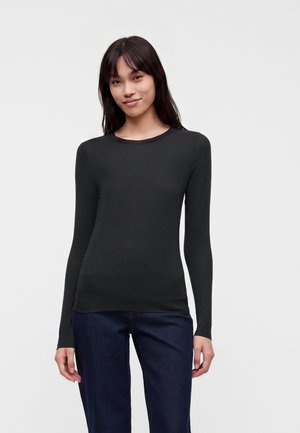 Jeune femme aux longs cheveux foncés portant un t-shirt noir moulant à manches longues et un jean bleu foncé, debout devant un fond clair uni.