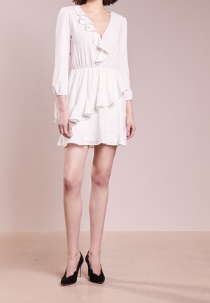 Femme portant une mini-robe blanche à volants avec des manches trois-quarts et des talons hauts noirs, debout devant un fond beige uni.