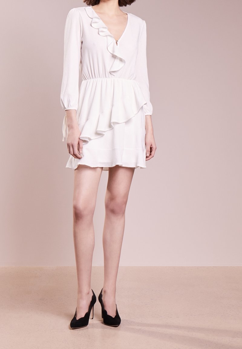 Femme portant une mini-robe blanche à volants avec des manches trois-quarts et des talons hauts noirs, debout devant un fond beige uni.