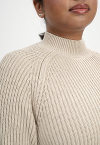 Beige Rippstrick-Pullover mit hohem Kragen, der strukturierte Linien und subtile Details an den Schultern aufweist. Glattes, warmes Material.
