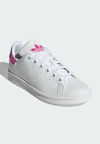 White leather sneaker with pink accents, textured surface, round toe, and flat laces. Features a small logo on the tongue and side.