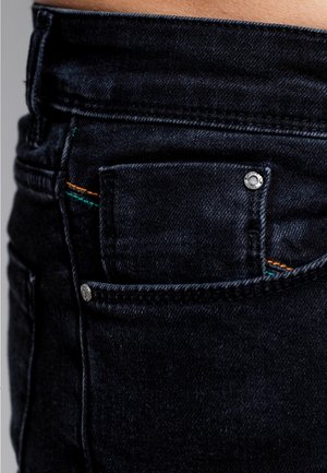 Vaqueros de mezclilla negra con un pequeño bolsillo delantero, herrajes plateados y detalles de costuras coloridas en naranja y verde. Textura suave.