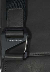 Detail einer schwarzen Ledertasche mit einem Metallverschluss, der mit "Piquadro" gebrandmarkt ist, genähten Kanten und einer strukturierten Oberfläche.